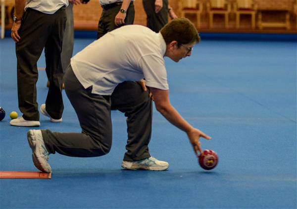 A bowler crouches to deliver a bowl on the blue indoor carpet at Newport Indoor Bowls Centre.