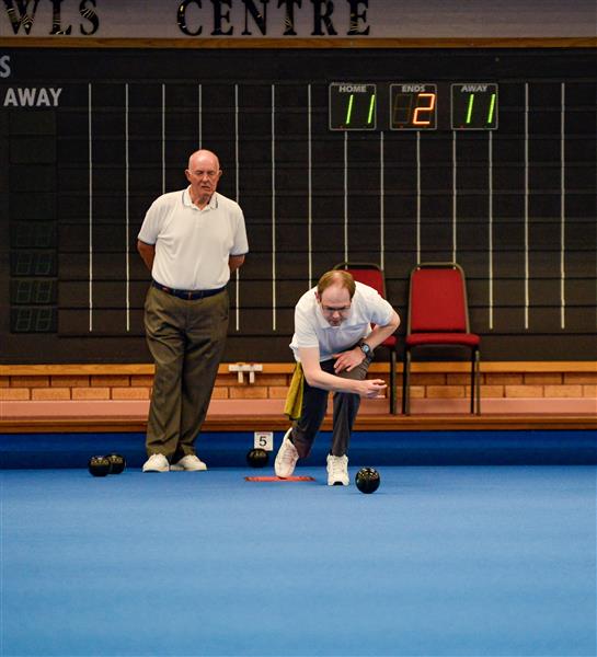 A bowler delivers in front of the scoreboard while a team-mate watches from the mat.